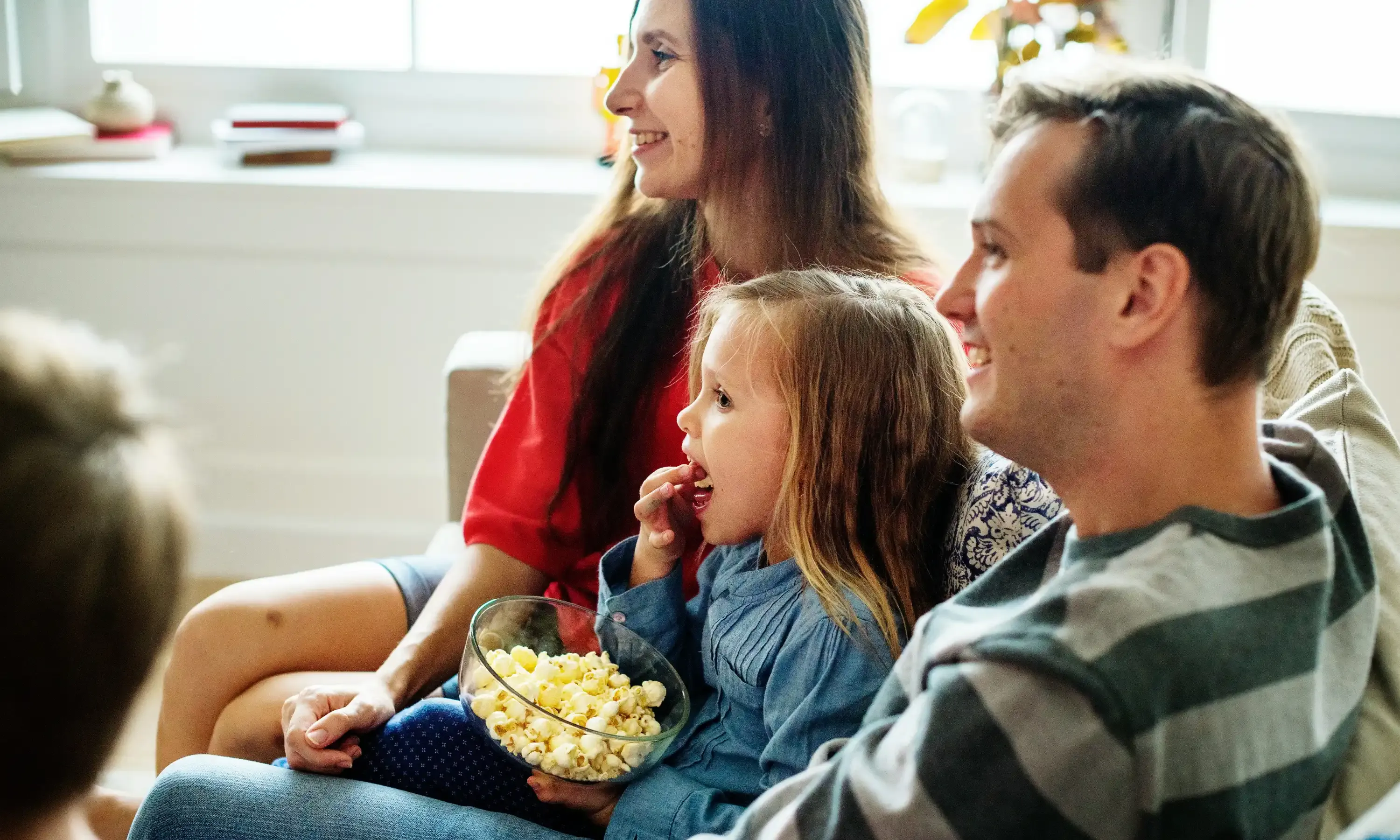 Familia viendo televisión en el sofá de su casa mientras una niña come palomitas